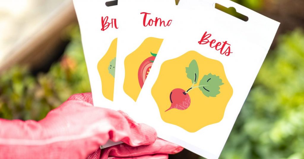 a hand wearing a red gardening glove holding three seed packets - beets, tomatoes, and broccoli